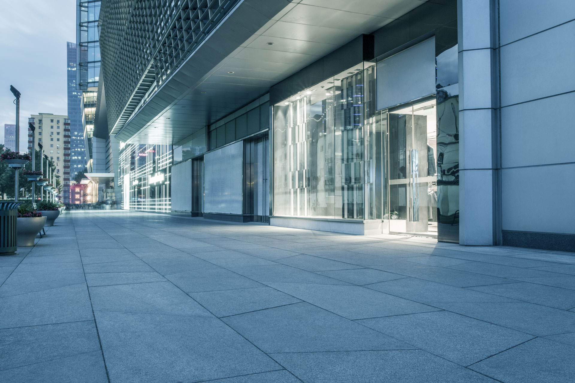 Retail storefront with lit glass windows along an empty commercial plaza at dusk.