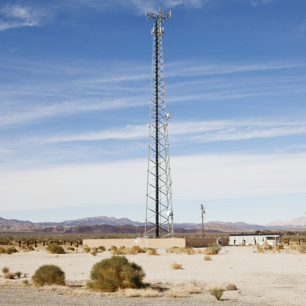 Cell tower with a fenced equipment compound in a remote desert landscape.