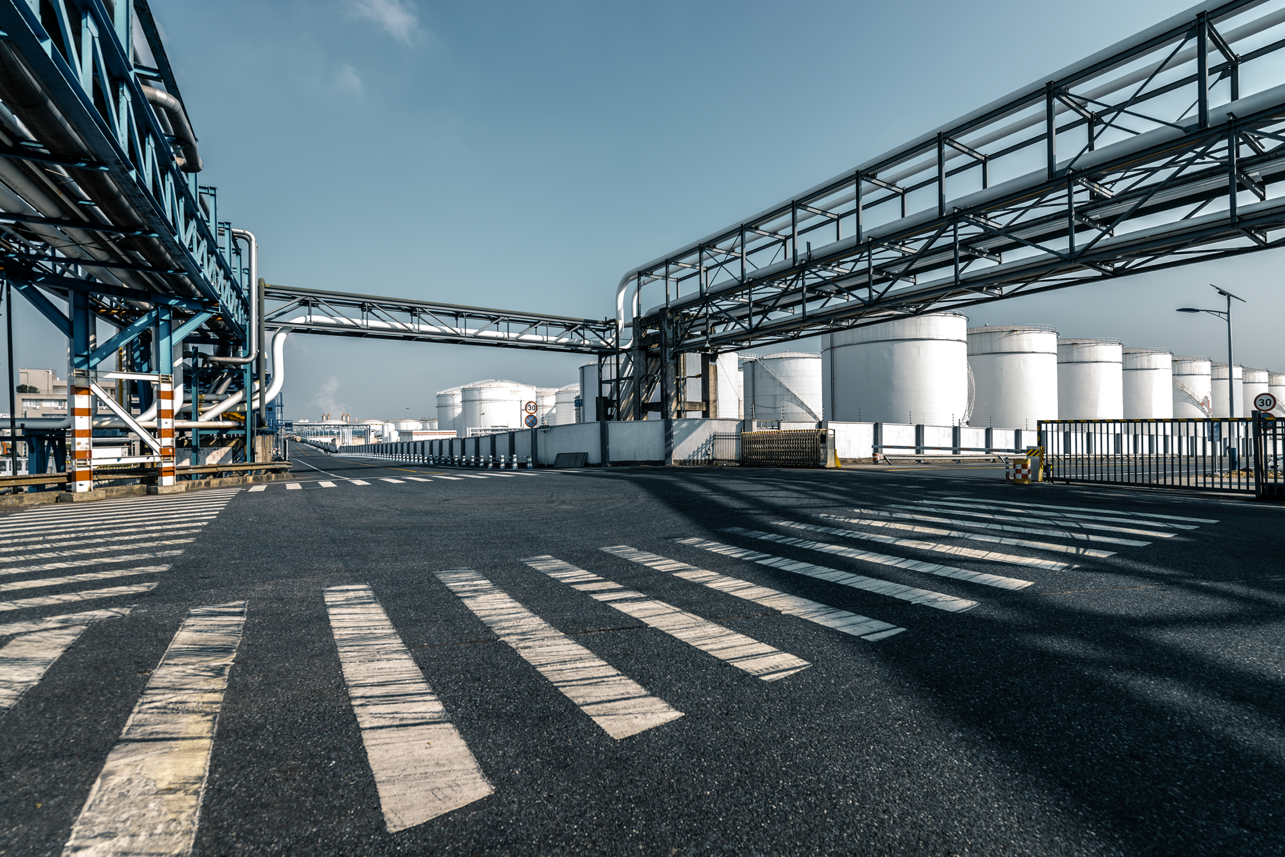 Industrial facility with large storage tanks, overhead piping, and an empty access road.